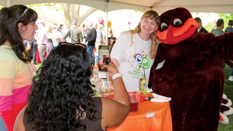 A student hugs and smiles with the HokieBird while another takes a photo with a phone under a tent.