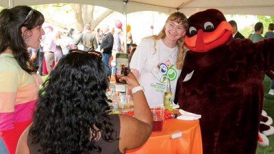 A student hugs and smiles with the HokieBird while another takes a photo with a phone under a tent.