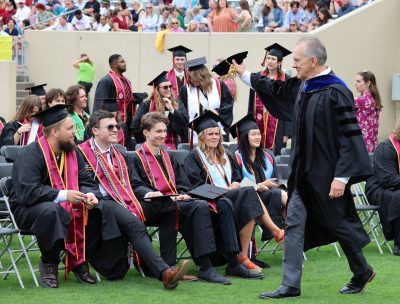 Paul Winistorfer walking on a football field dressed in regalia waves to students at commencement