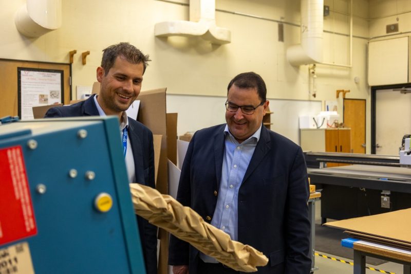 Two men in suits look at a machine that crinkles brown paper for packaging.