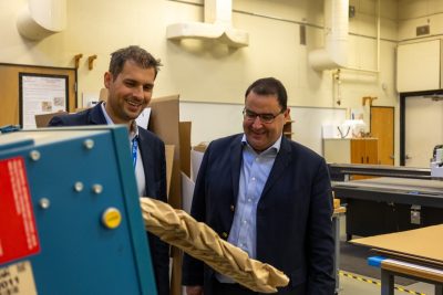 Two men in suits look at a machine that crinkles brown paper for packaging.
