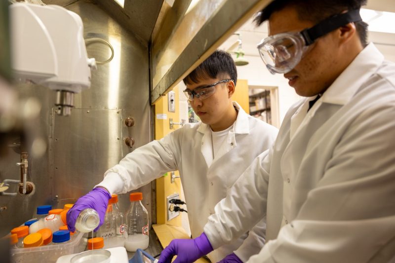 two students in white lab coats and eye protection in a lab