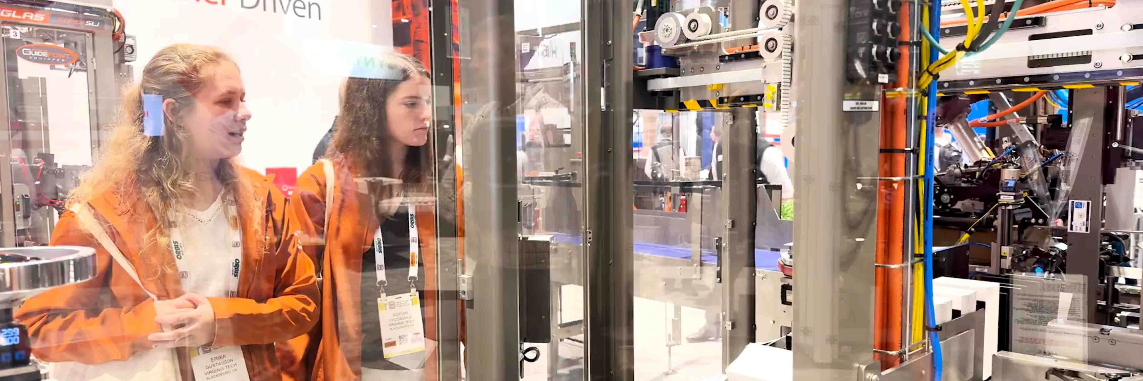 Two students look at a machine packaging assembly behind plexiglass