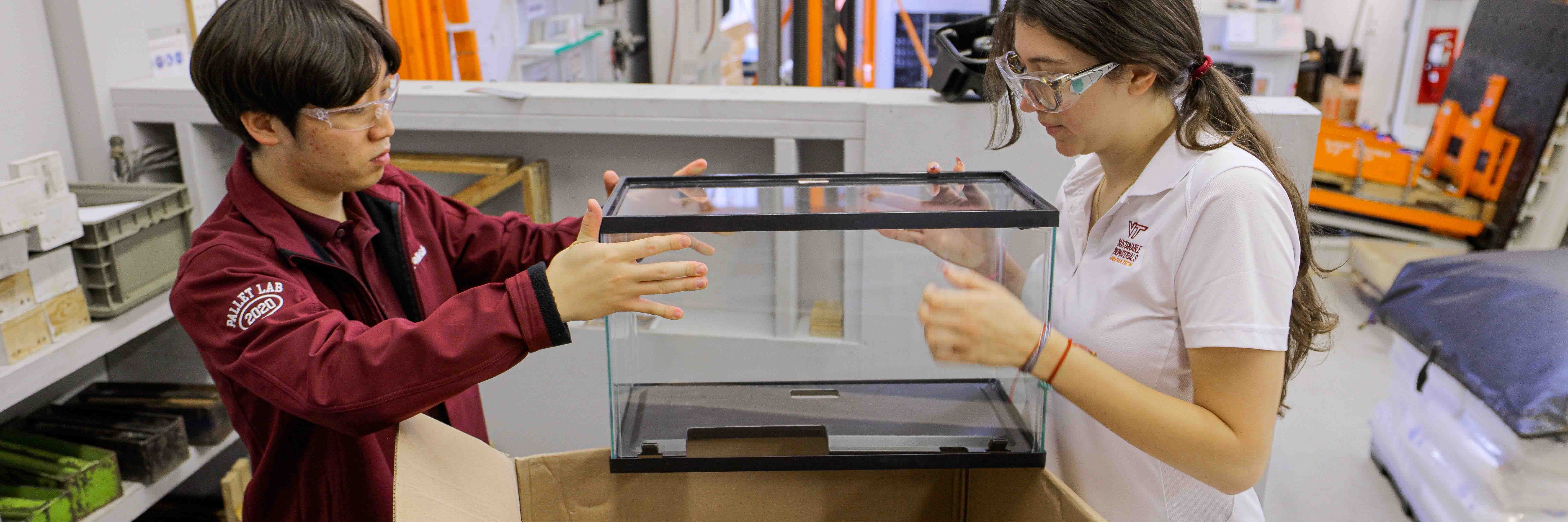 Two students lift a fish tank carefully into a corrugated cardboard box.