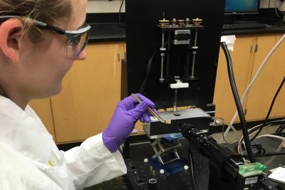 A student wearing a lab coat, protective eyewear, and latex gloves holding a sample of wood with tweezers near a machine.