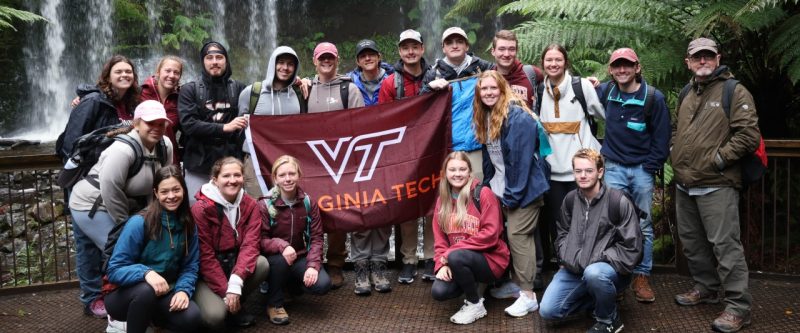 Students and a professor in front of a waterfall holding a flag with the Virginia Tech logo.