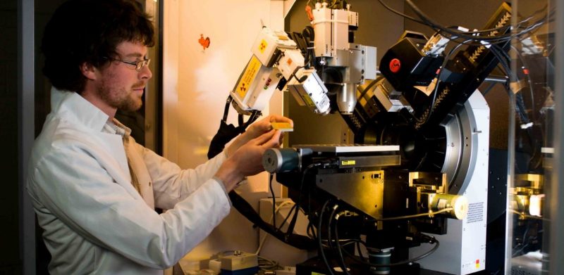 A graduate student wearing a white lab coat loading a wood sample onto an x-ray machine