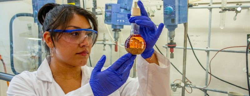 A student researcher in a lab holds a beaker with a liquid and solid up to eye level